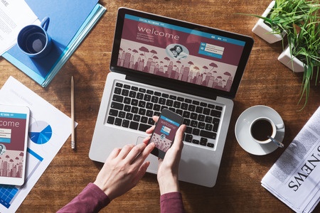 Laptop and cell phone on a table with coffee cups and documents. Woman's hands holding cell.