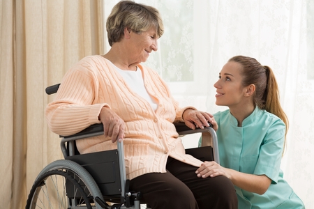 Young woman kneeling by older woman in wheelchair.