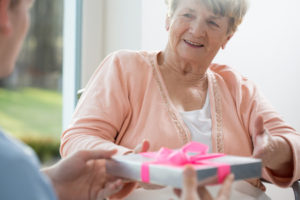 Woman in blue blouse giving gift to woman wearing pink sweater over white blouse, sitting in a wheelchair..