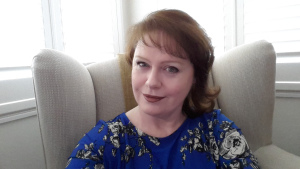 Sam Moss, smiling, sitting on a couch, wearing a blue patterned blouse.