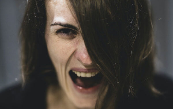 Woman with long black hair partially hanging over her face. Wearing a black shirt. Very unhappy. Looks like she's screaming.