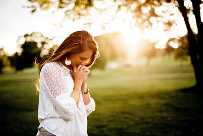 Woman, head bowed, hands under chin. Standing in green meadow with trees.
