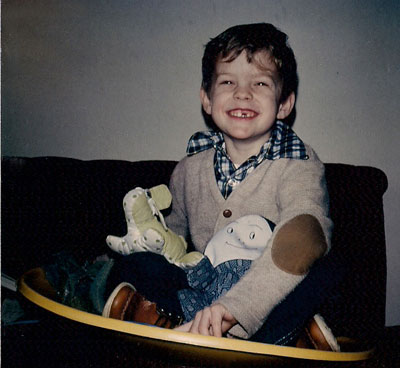 Bill sitting on a brown couch on a snowdish holdng his stuffed animals. He's smiling big, missing a front tooth.