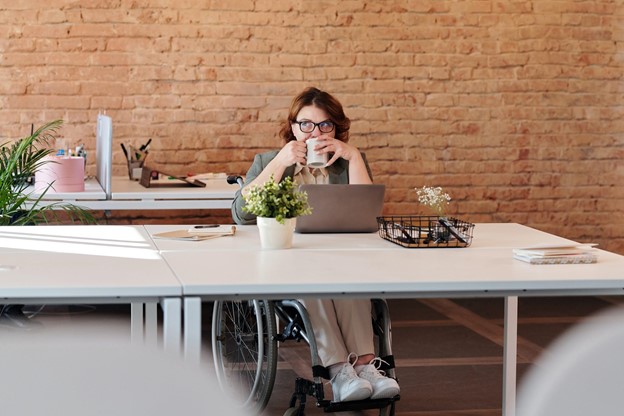Disabled woman in wheelchair, sipping coffee, sitting in front of her laptop, at her job.