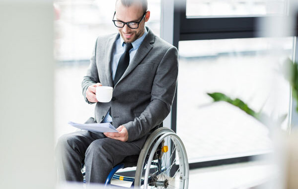Disabled man in business suit in wheelchair in an office, holding cup of coffee, reading a paper.