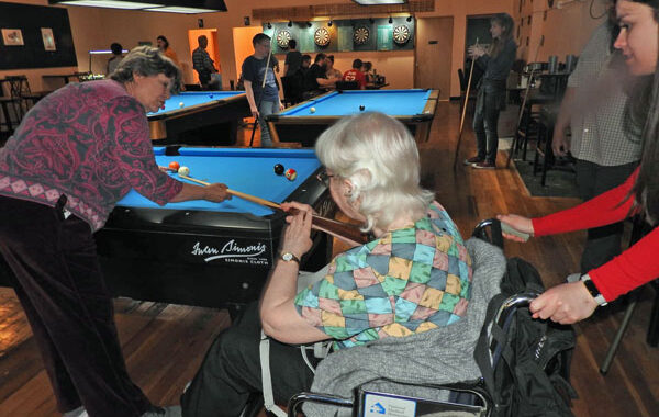 Carole using cue stick while seated in her wheelchair. Pushed by grand-neice, Elaina. Friend Abby helping her to shoot. At her family reunion.