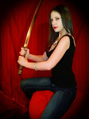 Jenny Jones dressed in a black jumsuit holding up a sword ready to slash. Redish background.