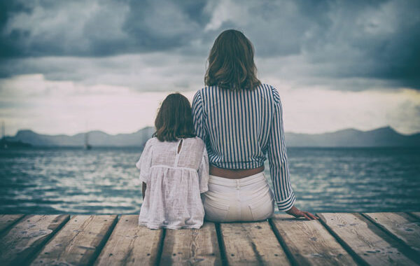 Mother and daughter sitting on wooden boardwalk watching from a distance a storm over a lake with mountains in background.